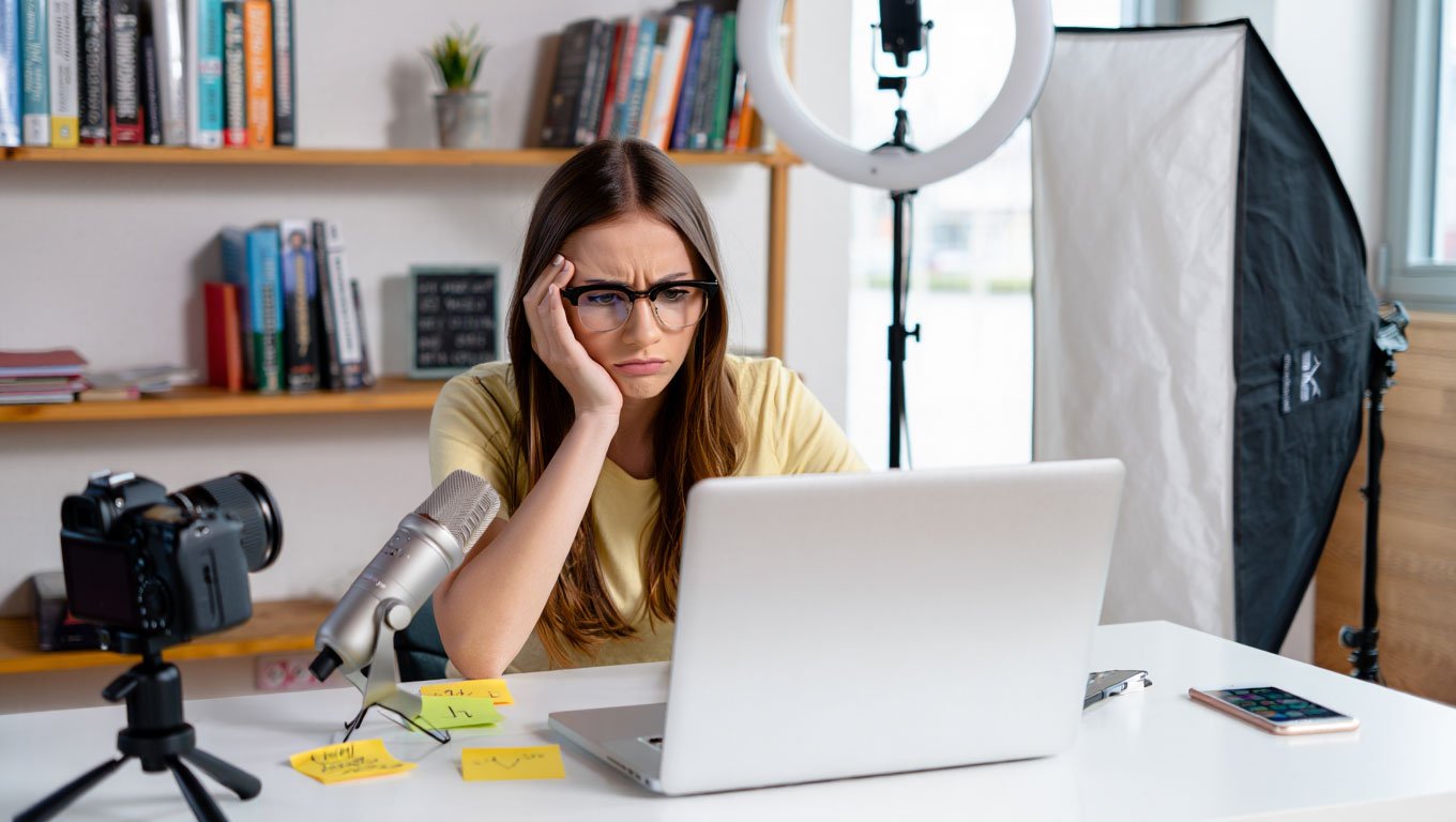 Mujer joven con expresión de frustración frente a su portátil en un estudio de grabación con cámara, micrófono y focos